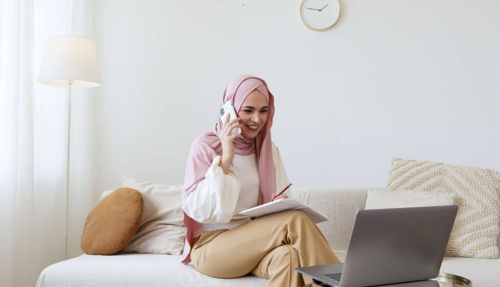 woman sitting on couch with notepad on her lap and cellphone to her ear. Laptop is on the table in front of her.