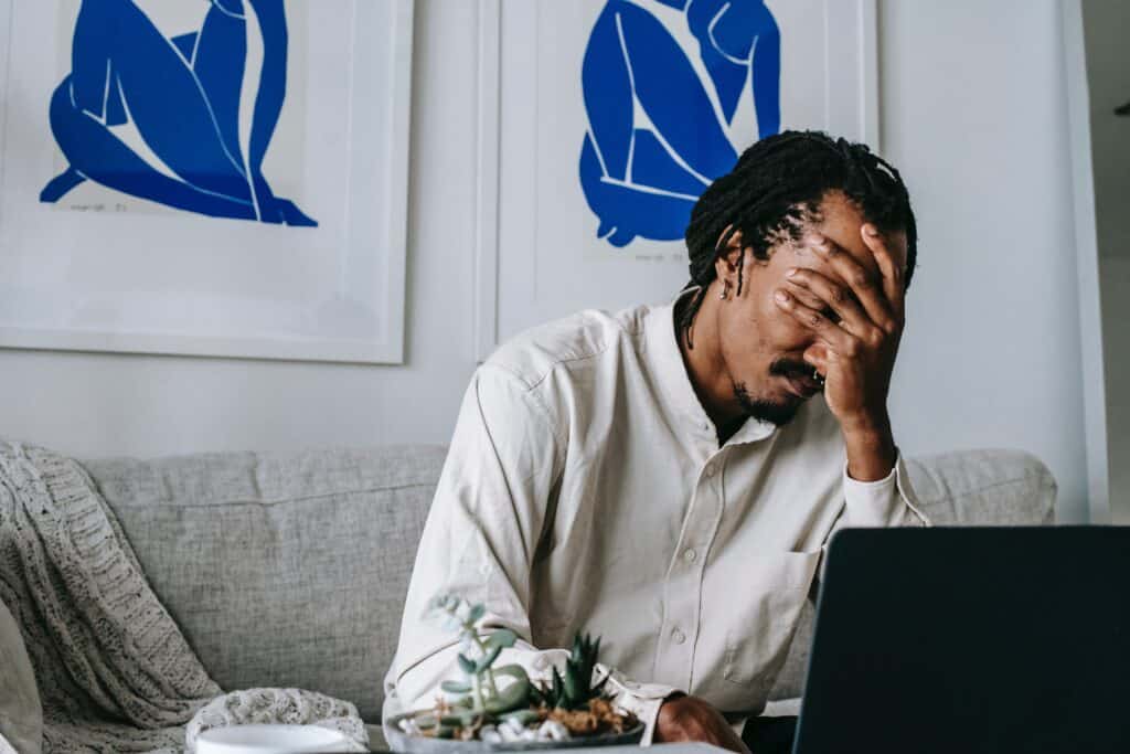 frustrated person with hand covering their facing sitting on couch in front of their laptop