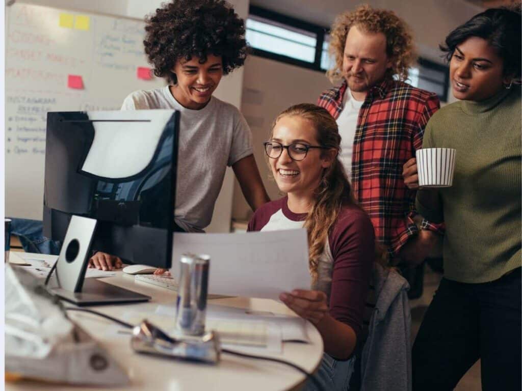 software engineers huddled around a desk where one of them is sitting. She is holding a piece of paper and showing it to the others.